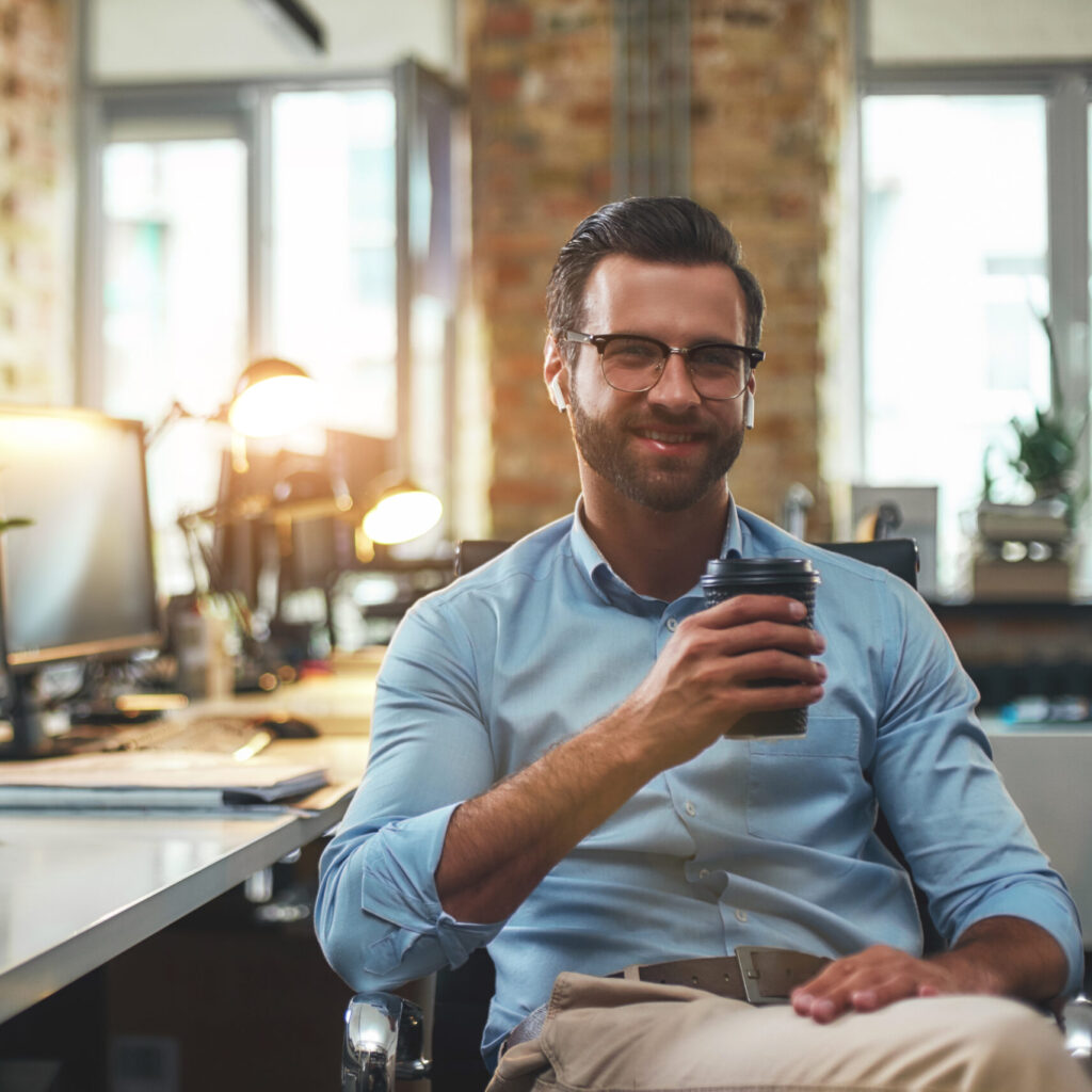 I can not work without coffee. Portrait of young bearded man in eyeglasses and headphones holding cup of coffee and smiling at camera while sitting in the modern office. Work concept. Business concept